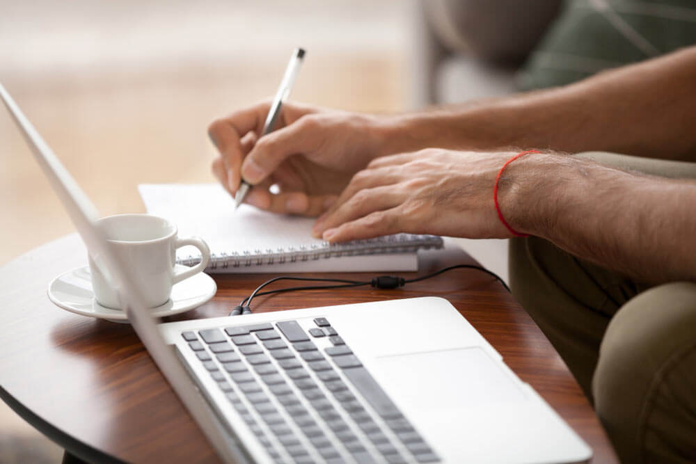 male use laptop writing down important data in notebook