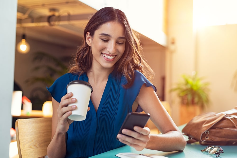 Young beautiful woman holding coffee paper cup and looking at smartphone