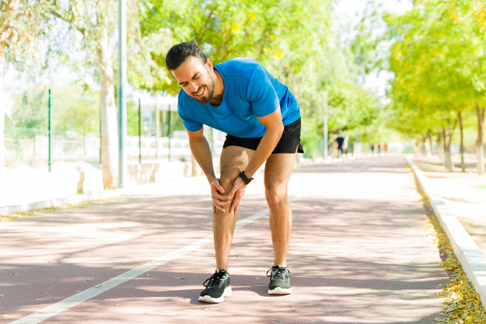 Hispanic sportsman feeling a sharp pain in his knee while exercising outdoors on a running track