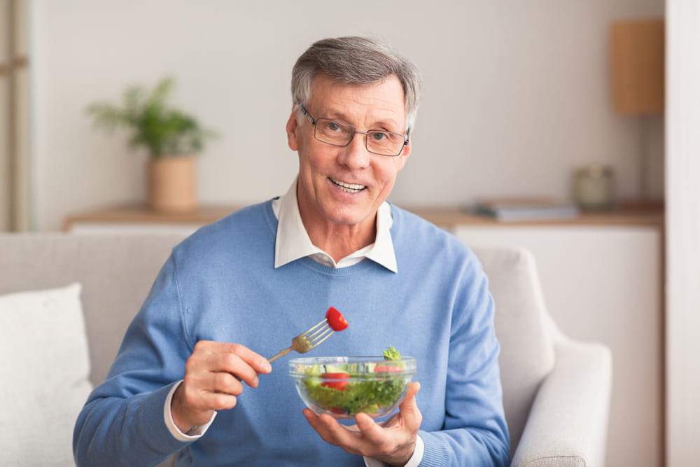 Healthy Nutrition. Senior Man Enjoying Vegetable Salad Smiling To Camera Sitting On Sofa At Home. Selective Focus