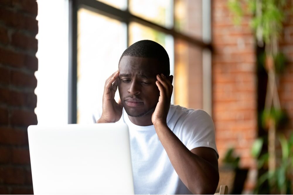 Boring black american millennial man sitting indoors looking at pc screen feels unwell