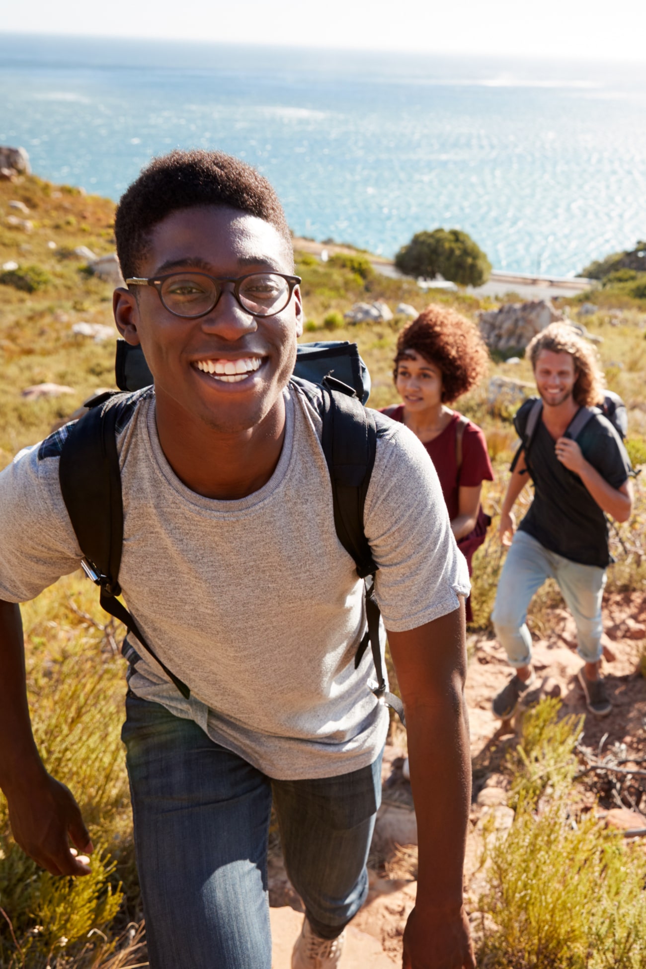 Millennial African American man leading friends hiking single file uphill on a path by the coast