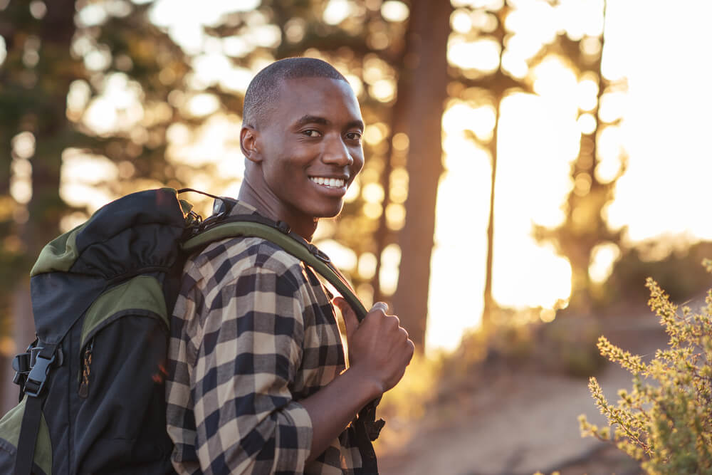Portrait of a smiling young African man wearing a backpack standing on a trail while hiking alone in the late afternoon