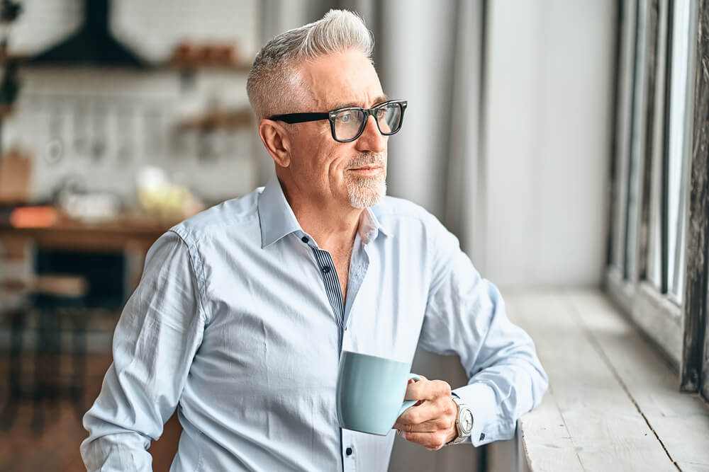 happy mature man in glasses looking out the window and holding cup