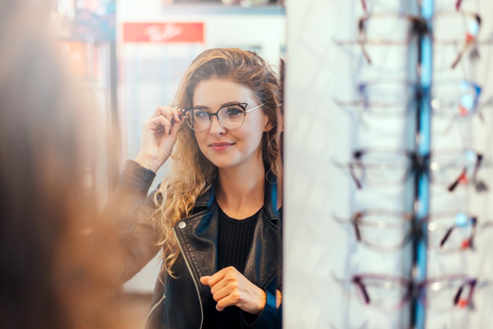 Smiling young woman trying on glasses on mirror in optician