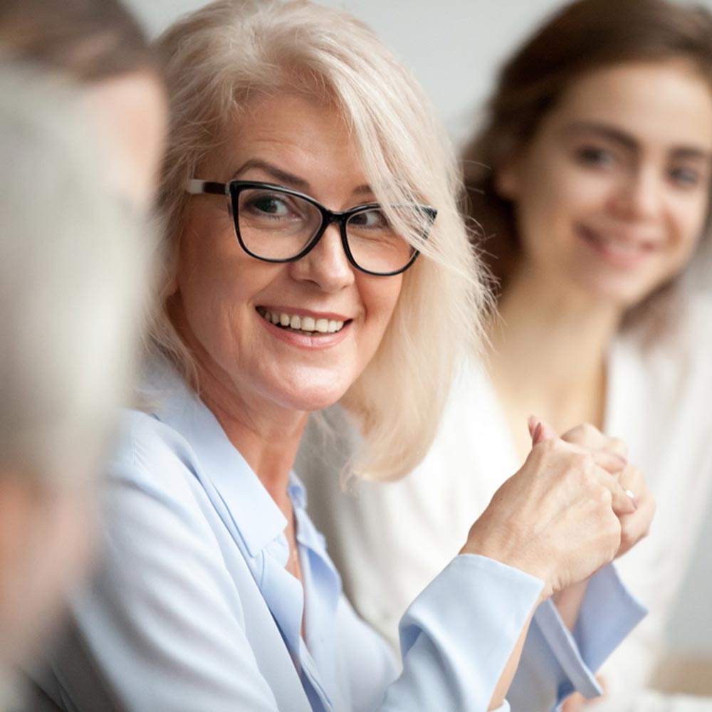 Smiling aged woman in glasses looking at colleague at team meeting