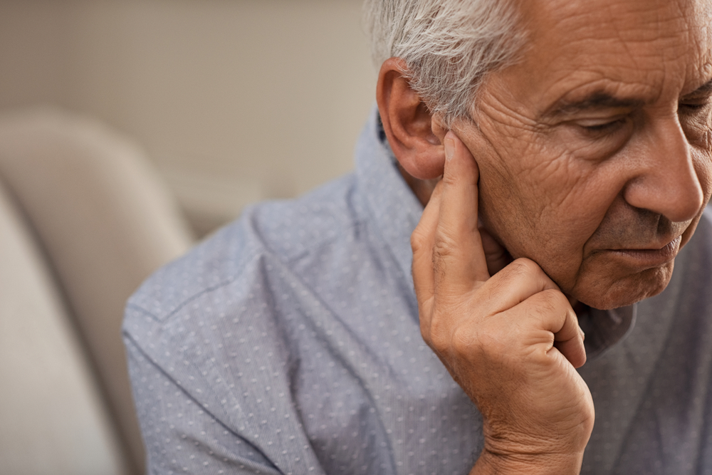 Mature man sitting on couch with fingers near ear suffering pain