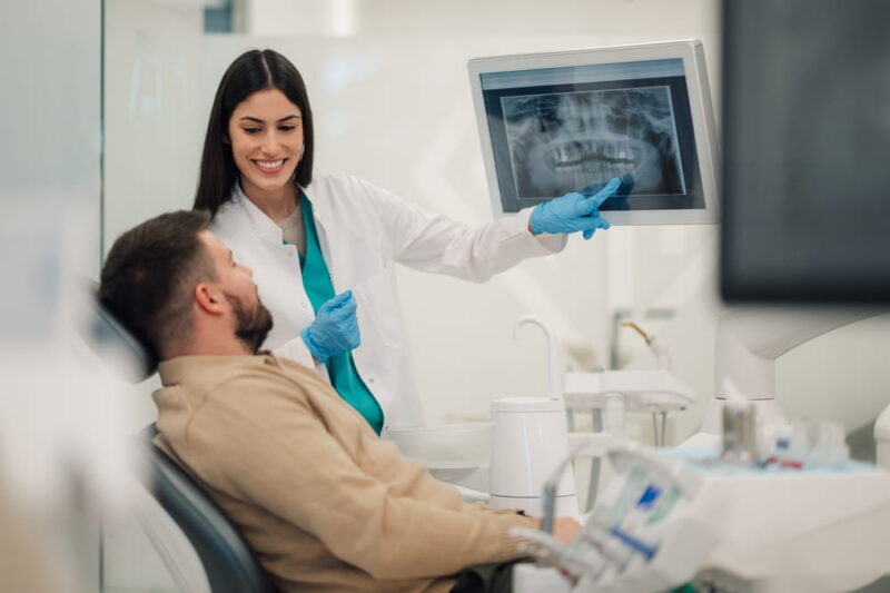 Smiling female dentist pointing at teeth x ray on digital screen, explaining diagnosis to male patient sitting in dental chair,