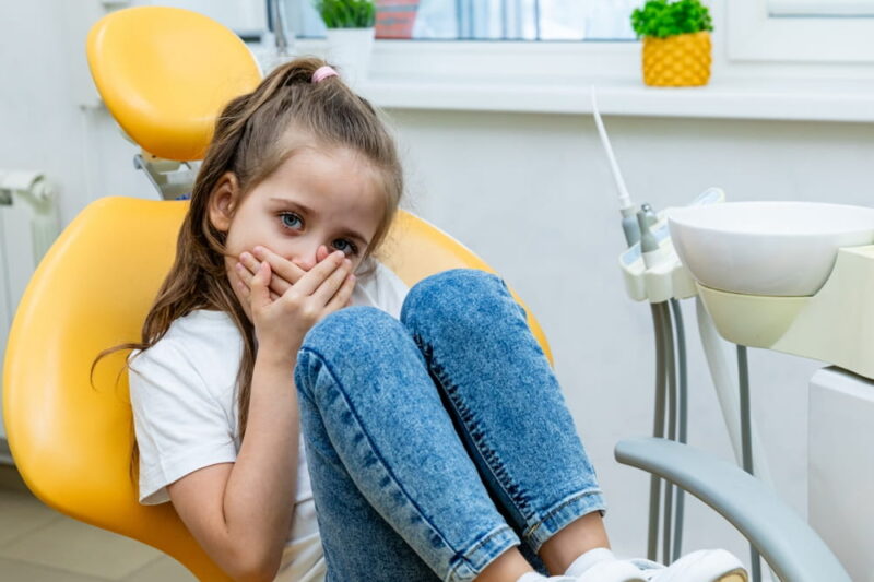 Childish fear. Cute little girl sitting in the dentist chair and covering her mouth in fear, being afraid of a dental treatment.