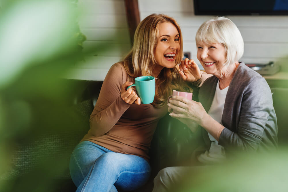 Happy senior mother with adult daughter sitting on couch and holding cups with coffee or tea at home.
