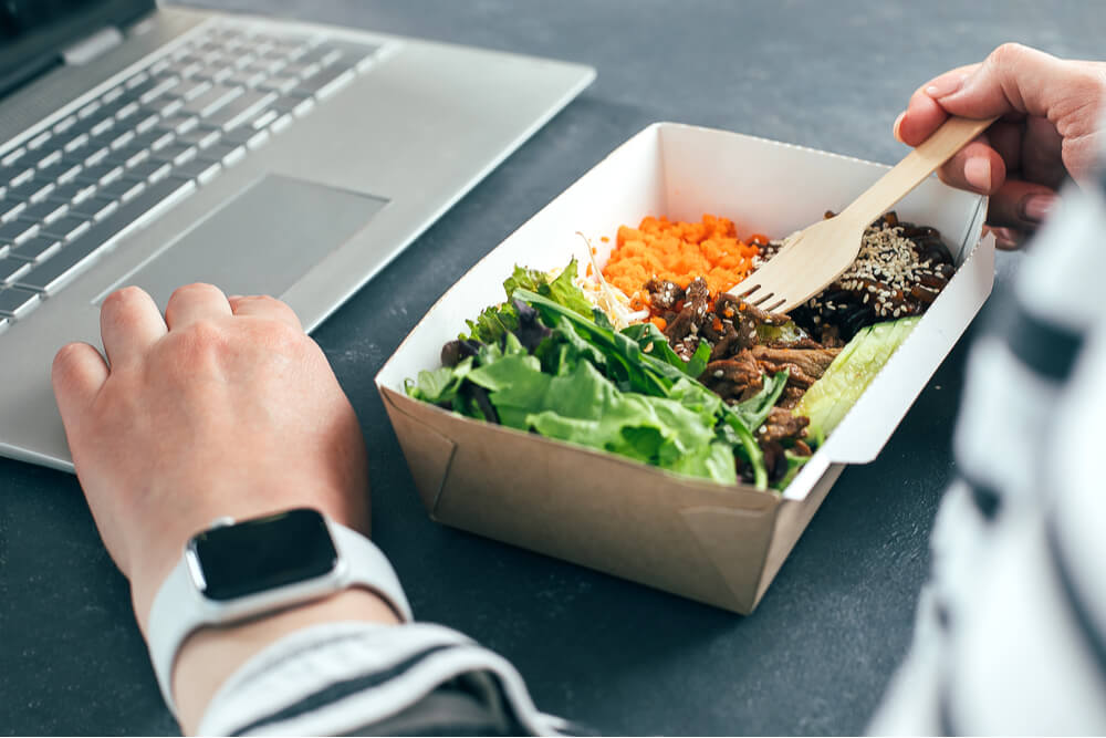 Woman having lunch from recycled bowl and using laptop. Concept of food delivery, quarantine, take out food