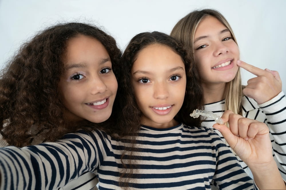 Three young beautiful multiracial kid girls make selfie holding an invisible braces aligner