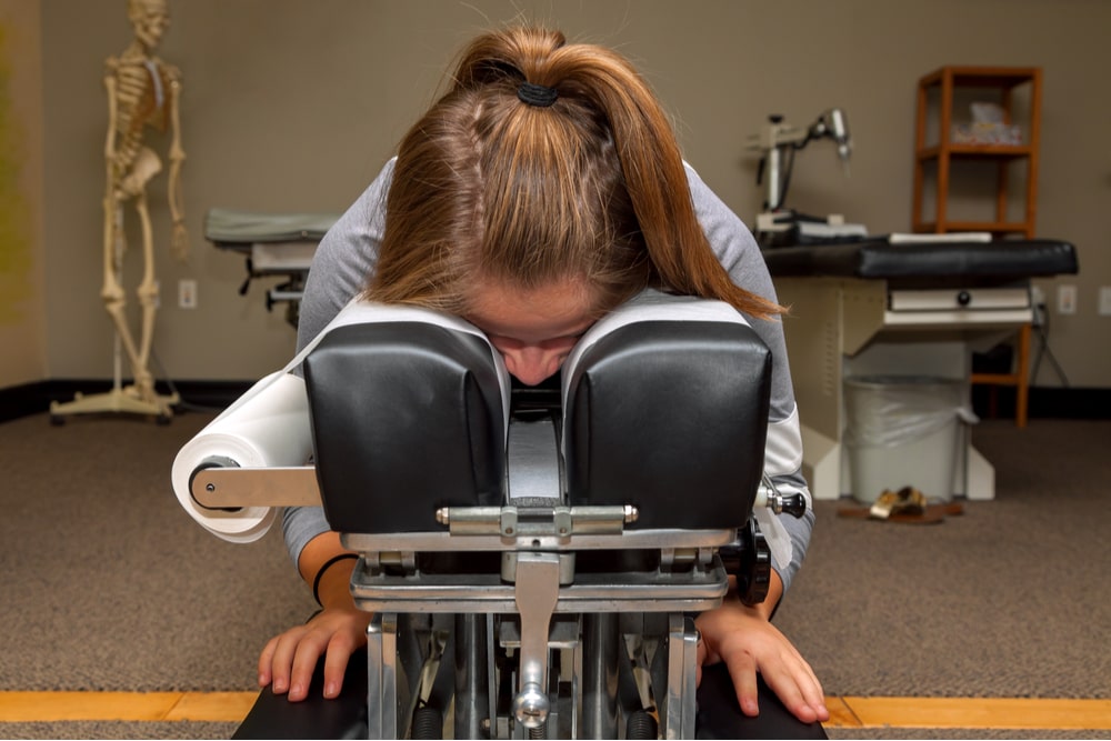 A young girl waits on a tilting chiropractic table with her face down in a crack in the table