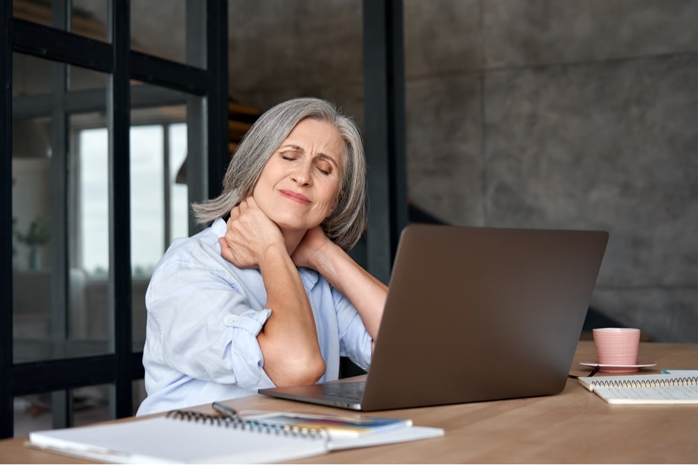 Tired stressed old mature business woman suffering from fibromyalgia neckpain working in office sitting at table